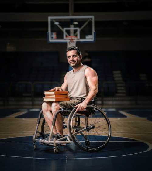 pessoa em cadeira de rodas com livros ao colo num campo de basquetebol